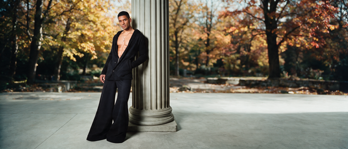 A man wearing a black wide-leg suit and an open black blazer stands leaning against a large stone column in an outdoor park setting with autumn trees.